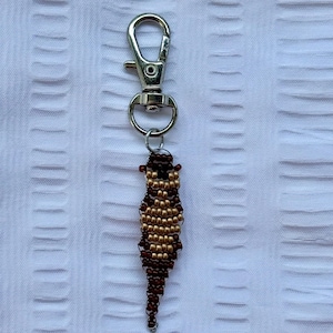 May include: A beaded keychain featuring a small, elongated otter design. The otter is crafted with brown and gold beads, attached to a silver-colored metal clasp. The keychain is a handmade accessory.