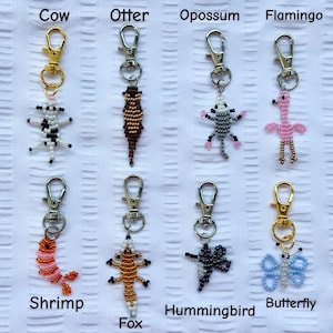 May include: A collection of beaded keychains featuring various animal designs, including a cow, otter, opossum, flamingo, shrimp, fox, hummingbird, and butterfly. Each keychain has a metal clasp for easy attachment. The keychains are made with colorful beads.