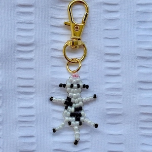 May include: A beaded cow keychain with a gold-colored clasp and ring. The cow is white with black spots, black hooves, and a pink flower on its head. The keychain is on a white textured background.