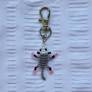 May include: A beaded opossum keychain with a silver clasp. The opossum is gray with black and pink accents. The keychain is made of small beads and is attached to a silver metal clasp.
