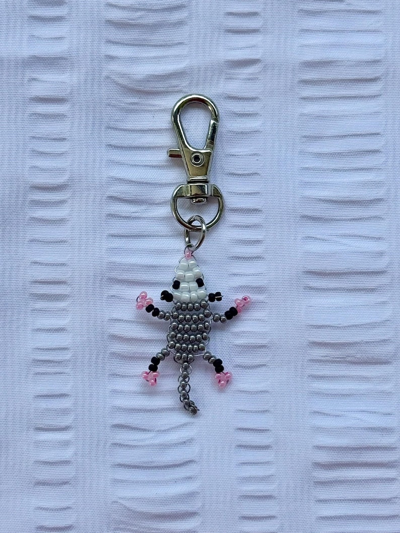 May include: A beaded opossum keychain with a silver clasp. The opossum is gray with black and pink accents. The keychain is made of small beads and is attached to a silver metal clasp.