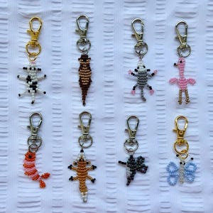 May include: A collection of beaded keychains featuring various animal and insect designs. The keychains include a cow, lizard, possum, flamingo, shrimp, gecko, dragonfly, and butterfly. Each keychain has a silver or gold-colored clasp.