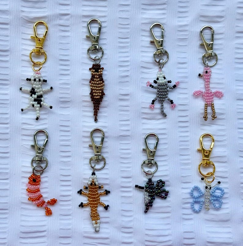 May include: A collection of beaded keychains featuring various animal and insect designs. The keychains include a cow, lizard, possum, flamingo, shrimp, gecko, dragonfly, and butterfly. Each keychain has a silver or gold-colored clasp.
