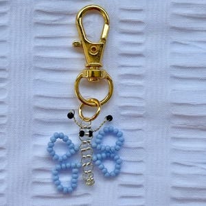 May include: A gold-colored metal keychain with a light blue beaded butterfly charm. The butterfly has a silver-colored wire body and black bead antennae. The keychain clasp is a lobster claw style.