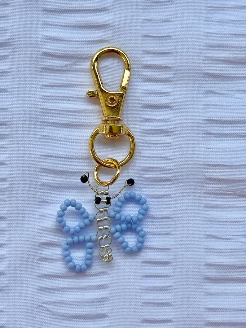 May include: A gold-colored metal keychain with a light blue beaded butterfly charm. The butterfly has a silver-colored wire body and black bead antennae. The keychain clasp is a lobster claw style.