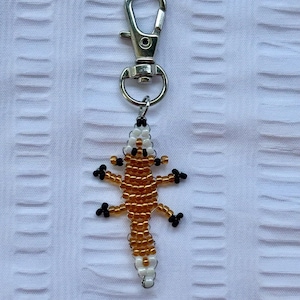 May include: A beaded lizard keychain with a silver clasp. The lizard is crafted with gold, white, and black beads, showcasing a detailed reptile design. The keychain is a decorative accessory.