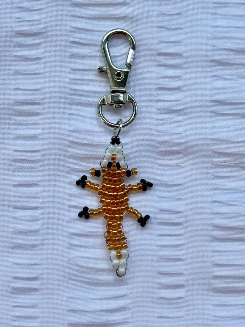 May include: A beaded lizard keychain with a silver clasp. The lizard is crafted with gold, white, and black beads, showcasing a detailed reptile design. The keychain is a decorative accessory.
