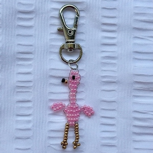 May include: A pink beaded flamingo keychain with gold-colored legs and a black beak. The flamingo is attached to a silver-colored metal clasp. The background is a white textured fabric.