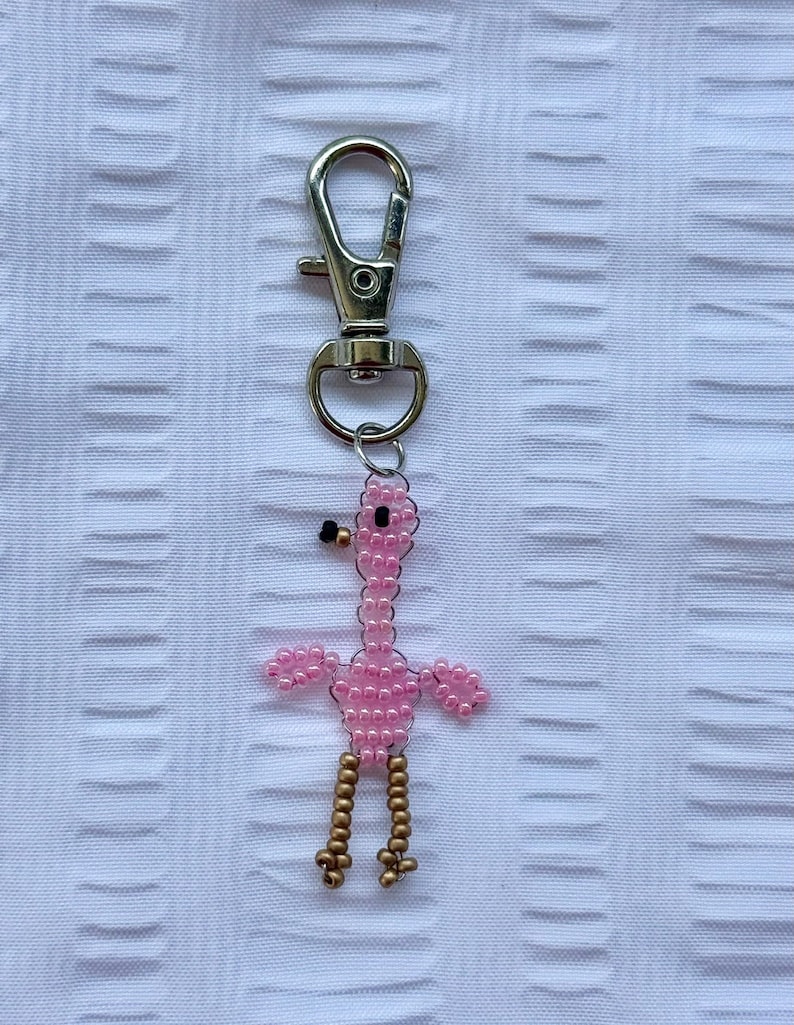 May include: A pink beaded flamingo keychain with gold-colored legs and a black beak. The flamingo is attached to a silver-colored metal clasp. The background is a white textured fabric.