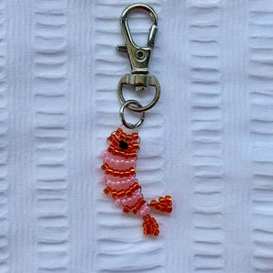 May include: A beaded keychain featuring a shrimp design. The shrimp is crafted with pink and orange beads, and has a black bead for the eye. The keychain has a silver-colored clasp.