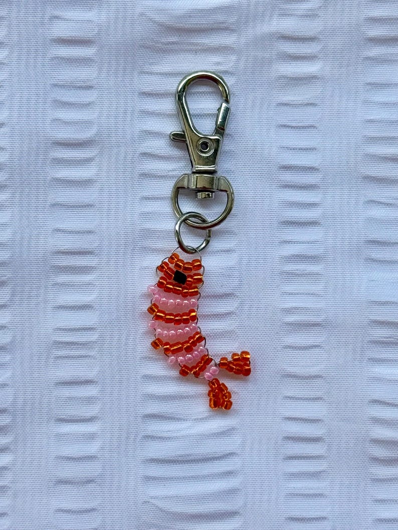 May include: A beaded keychain featuring a shrimp design. The shrimp is crafted with pink and orange beads, and has a black bead for the eye. The keychain has a silver-colored clasp.