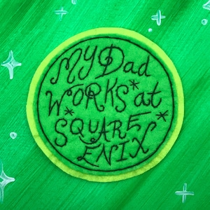 Peut inclure: Un patch en feutre vert avec une bordure jaune et le texte "My Dad Works at Square Enix" en écriture cursive noire.