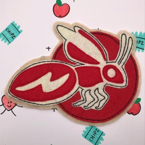 May include: A red and white felt applique in the shape of a rabbit-like creature with a lightning bolt design. The applique is on a beige background with red apples, orange, and the word "CHIPZ" on a teal rectangle.