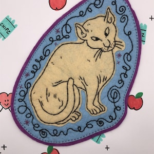 May include: A felt cat applique in a light yellow color, centered on a blue oval with a purple border. The cat is outlined in black thread, with embroidered details. The background includes drawn apples, a heart, and a snack package.