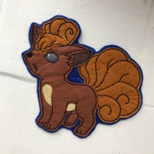 May include: A brown and orange felt appliqué of a Pokémon character with a curled tail. The character has a brown body, orange hair, and a blue eye.