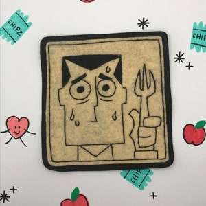 May include: A felt illustration of a cartoon character with a worried expression holding a fork. The character is framed in a brown rectangle with a black border. The background is white with red apples, a heart with legs, and blue and green chip bags.