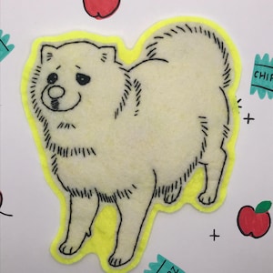 May include: A handmade felt dog applique with a yellow outline, black details, and a smiling face. The design includes red apple and "CHIPZ" illustrations on a white background. The dog is cream-colored.