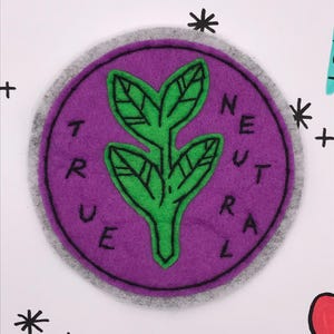 May include: A round, purple felt patch with the words "TRUE NEUTRAL" in black lettering. A green plant design is in the center. The patch has a gray border and is on a white background with additional doodles.