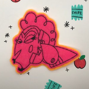 May include: A pink felt applique of a stylized face with an orange outline and black details. The image includes two red apples with green leaves, two teal rectangles with the word "CHIPZ", and black plus and star shapes on a white background.
