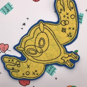 May include: A yellow felt bird-shaped applique with a blue border, featuring black line art and decorative markings. The background includes drawn apples, hearts, and the text "CHIPZ" on a teal banner.