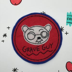 May include: A red felt patch with a blue border. The patch features a white cartoon bear wearing glasses and a mustache. The text "GRAVE GUY" is embroidered in black thread below the bear.