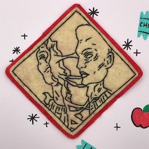 以下が含まれることがあります： 赤い縁とクリーム色の中心部を持つフェルトパッチ。中央には2つの顔の黒い線画があります。背景には、描かれたリンゴ、アスタリスク、プラス記号、ターコイズ色の「CHIPZ」ラベルが含まれています。