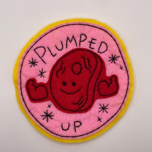 May include: A circular felt patch featuring a pink background with a yellow border. In the center is a red, smiling, muscular steak character with flexed arms. The text "PLUMPED UP" is embroidered in black, surrounded by small black star accents.
