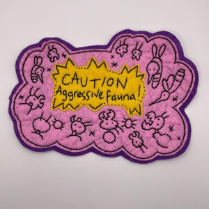 May include: A pink felt patch with a purple border featuring a yellow jagged speech bubble in the center with the black embroidered text CAUTION Aggressive Fauna! surrounded by black stitched illustrations of various small insect-like creatures.