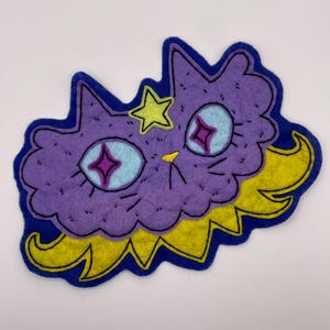 May include: A purple felt cat-shaped patch with a dark blue border. The cat has light blue eyes with purple star-shaped pupils, a yellow star on its forehead, a small yellow nose, and a yellow jagged collar-like shape at the bottom.