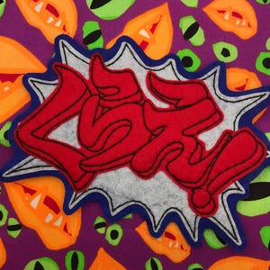 May include: A purple fabric with a repeating pattern of orange and green shapes, and green eye shapes. A red felt applique with the word "LOVE" in a graffiti style is centered on the fabric, outlined in blue and white.