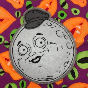 May include: A felt moon-shaped patch with a smiling face and a gray cap. The patch is gray with black details and a gray border. The background is purple with orange and green eye and mouth shapes.
