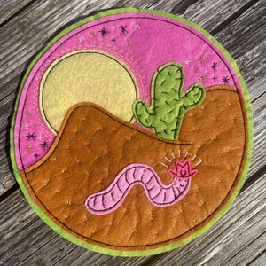 May include: A round felt coaster featuring a desert scene. The design includes a yellow sun, a green cactus, and a brown landscape. A pink worm wearing a cowboy hat is in the foreground. The coaster has a pink and green border, with embroidered stars.