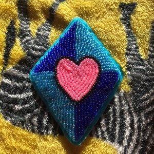 May include: A diamond-shaped beaded heart ornament with a pink heart in the centre. The ornament is made with blue and turquoise beads.