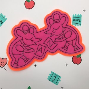 May include: A pink felt patch with an orange outline featuring two cartoon mice carrying a large letter A. The patch is surrounded by drawings of apples, oranges, and the word "CHIPZ" in a turquoise box.