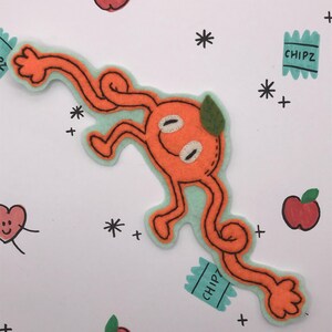 May include: Orange felt creature with a smiling face, arms, and legs, outlined in light green. The background features drawn apples, stars, and small bags of "CHIPZ". The design is whimsical and playful.