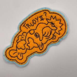 May include: An orange felt patch with a light blue border featuring black embroidered outlines of a fish and the text TRUDY II? with a starburst shape. The design includes a fish with a circular eye and a tail fin, all rendered in thread.