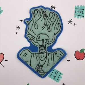 May include: A teal felt patch with a blue border, depicting a stylized figure with flame-like hair. The design includes black stitched details and is surrounded by drawn apples, asterisks, plus signs, and a "CHIPZ" tag.