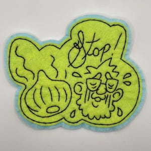 May include: A lime green felt patch with a light blue border featuring black embroidered outlines of an onion and a person wearing glasses with the word Stop! written above. The design includes small teardrop shapes around the person's face.