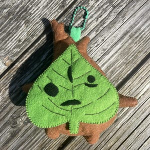 May include: A handmade felt Korok character plushie. The plushie features a green leaf face with stitched details and a brown body. A green beaded loop is attached for hanging. The plushie is designed to resemble a character from a video game.