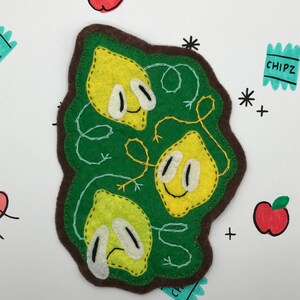 May include: Three felt lemon-shaped characters with faces, two smiling and one with a sad expression, are stitched together on a green felt background. The background is surrounded by a brown felt border.