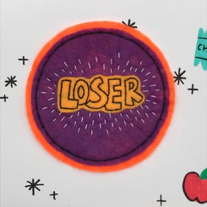 May include: A purple and orange felt circle with the word "LOSER" embroidered in yellow on it.