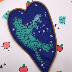 May include: A heart-shaped patch with a teal cat holding a blue fish against a dark blue background. The patch is outlined in brown and decorated with white stars. The background has illustrations of fruit and snack packaging.