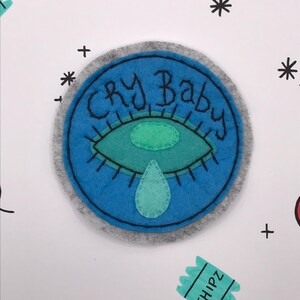 May include: A round, blue felt patch with the words "CRY BABY" in black script. The patch features a teal eye with a teardrop, all on a gray background. Decorative elements include black asterisks, plus signs, and a small teal chip packet.