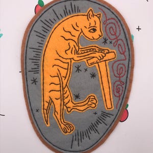 May include: An orange cat-like figure with black outlines, playing a wooden instrument, is centered on a gray oval patch with a brown border. The patch is decorated with black lines and stars. The background is white with small black crosses and cartoon fruit.