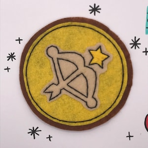 May include: A felt patch featuring a Sagittarius symbol: a bow and arrow with a star. The patch has a yellow centre with a brown border. Black asterisks and plus signs surround the patch on a white background.