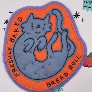 May include: A handmade felt cat-shaped patch in blue, orange, and purple. The cat is lying on its back with the words "FRESHLY BAKED BREAD ROLL" in black lettering around the edge. The patch is surrounded by decorative elements.
