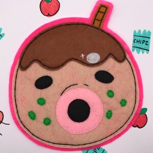 May include: A felt coaster in the shape of a cartoon character's face, featuring a pink outline, brown hair, and a pink mouth. The coaster has black eyes, green spots, and a small silver star. The background includes apple and "CHIPZ" graphics.