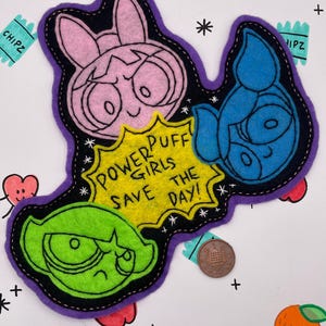 May include: A felt patch featuring the Powerpuff Girls with a pink, blue, and green character head, and a yellow speech bubble with the text "POWERPUFF GIRLS SAVE THE DAY!" in black. A copper coin is placed next to the patch for scale reference.