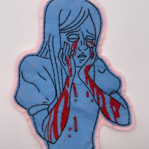 May include: A light blue felt patch featuring an embroidered illustration of a person with long hair, hands pressed to their face, and red beaded tears and blood-like streaks running down their cheeks and arms, outlined in black with a pink border.
