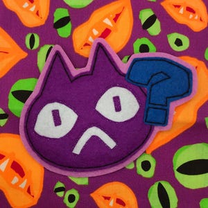 May include: A purple cat-shaped felt patch with white eyes and a blue question mark, set against a purple background with orange and green monster eye and mouth designs. The cat has a worried expression.